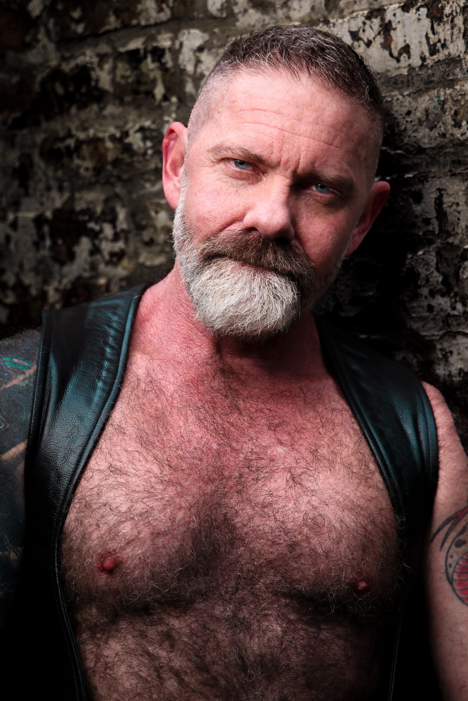 Leather portrait: a silver-bearded, broad-chested man in a leather vest holds the lens with a steady gaze against a weathered stone wall.