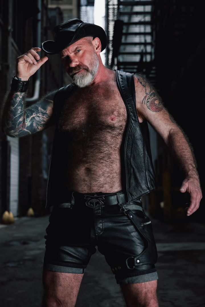 A silver-bearded man in a leather vest, cap, and shorts tips his cap brim in a dim San Francisco alley, a fire escape rising behind him.