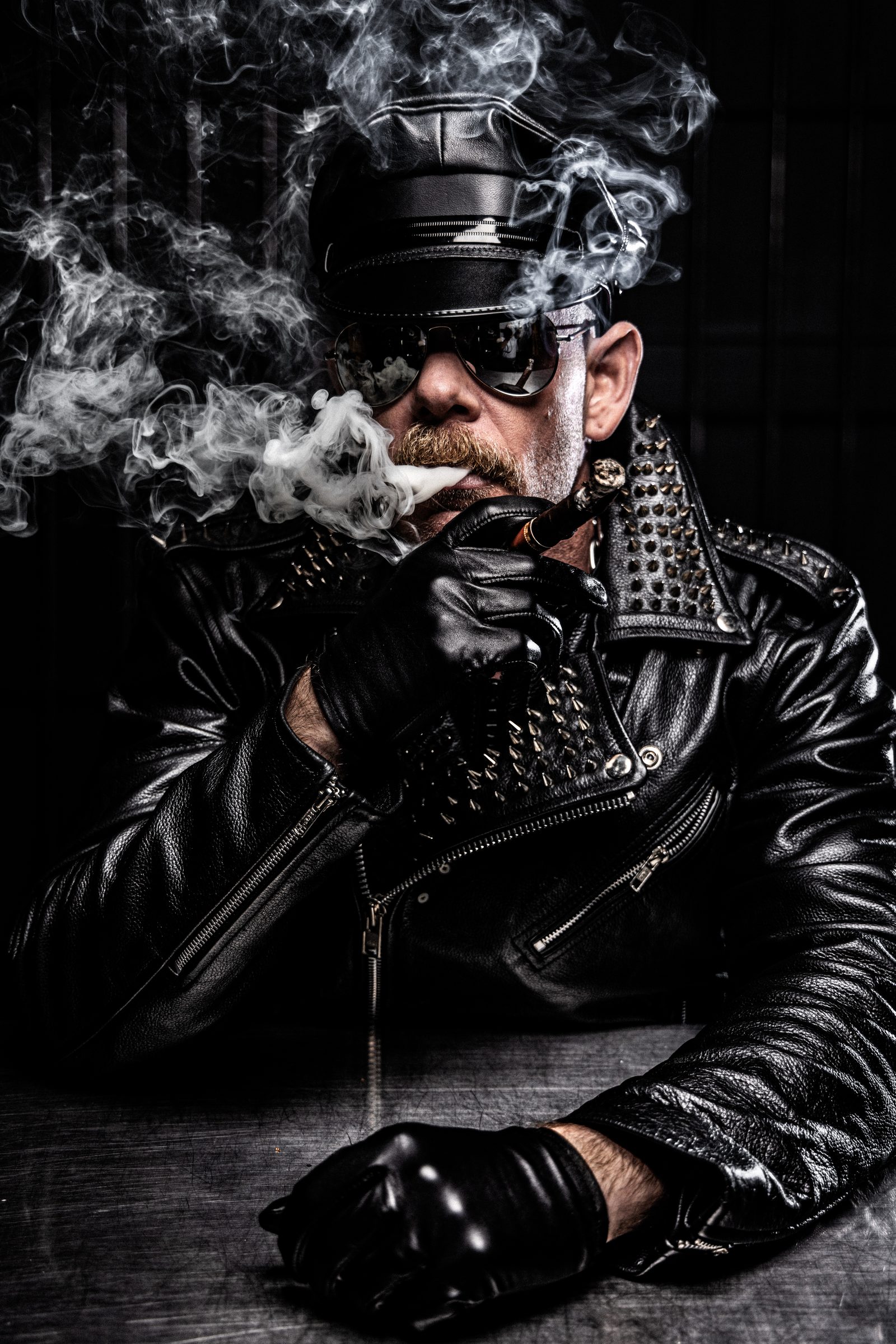 Leather portrait: an older, broader man in a cap and jacket exhales cigar smoke, sculpted from near-black by a single hard strobe.