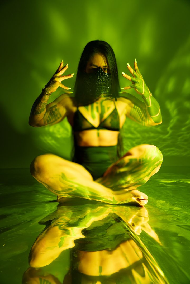 A cross-legged figure lit chartreuse and gold, projected light falling across the body on a dark ground.