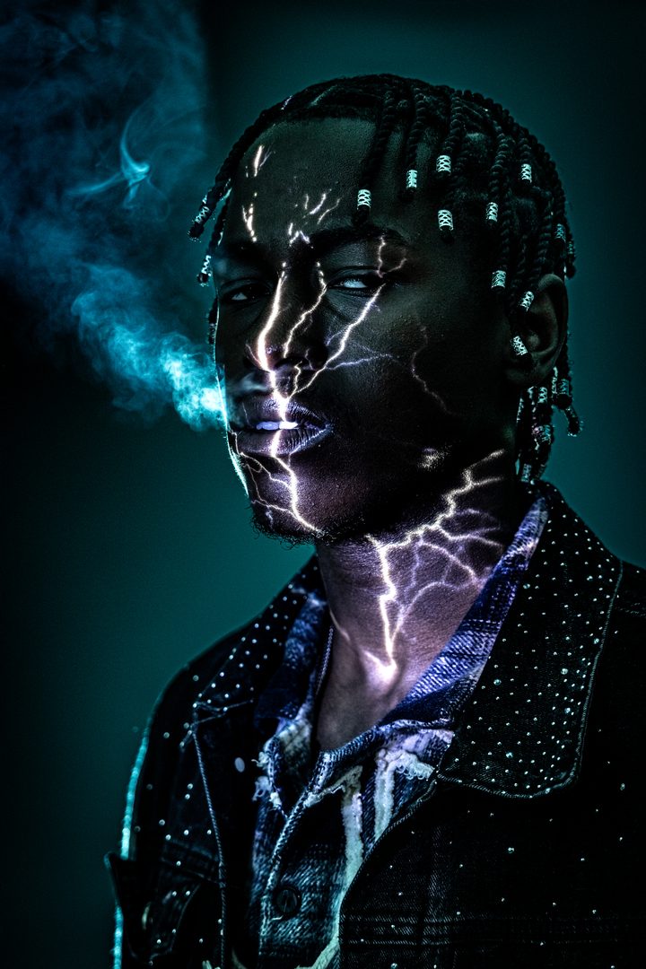 Lightning projected across a man's face and neck on a teal ground, smoke crossing the frame, the figure sculpted from black.