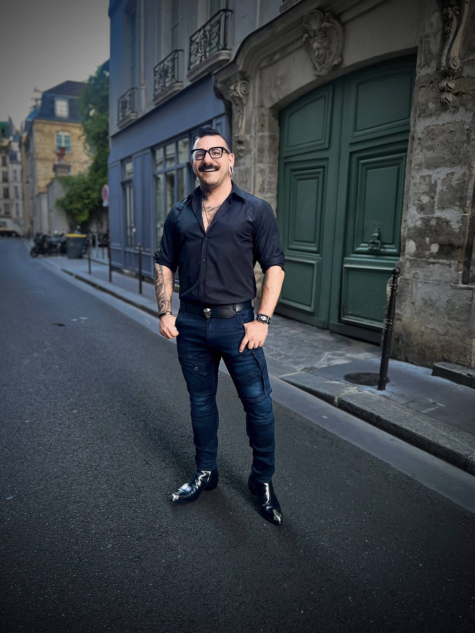 Nick Leoni, the photographer behind Nick Leoni Studios: a smiling man with dark-framed glasses and a styled mustache, in an open black shirt and dark jeans, standing on a quiet Paris street.